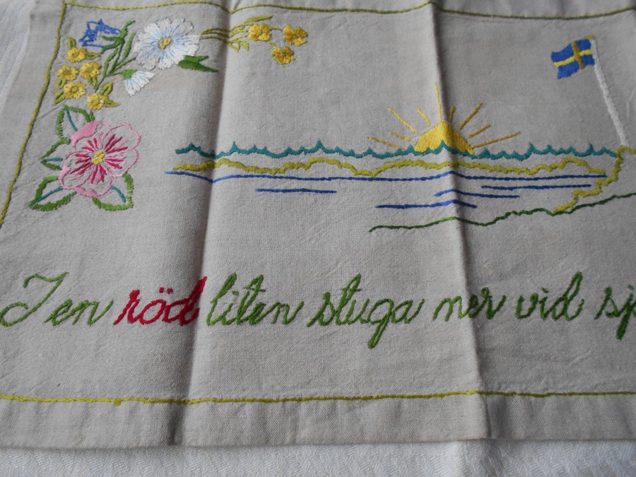 Broderad tavla med motiv av en stuga vid sjön