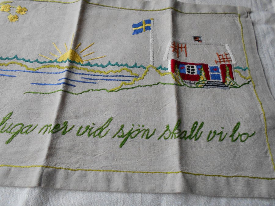 Broderad tavla med motiv av en stuga vid sjön